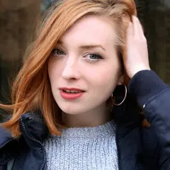Sandra Beijer, med sitt röda hår och ljusa ögon, ler lätt medan hon rör vid sitt hår. Hon bär en blå jacka och en ljusblå stickad tröja.