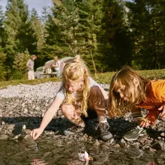 Två barn leker vid en sjö med små båtar, som en del av sina femkamp lekar. I bakgrunden syns ett tält och träd.