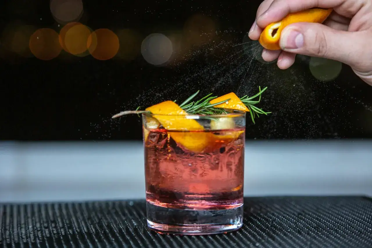 En hand pressar en apelsin över en Aperol Spritz, som garneras med apelsinskal och rosmarin.